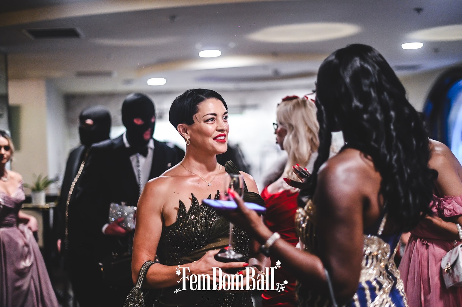 Two dommes chatting at the femdom ball, in the background two submissive men serving drinks